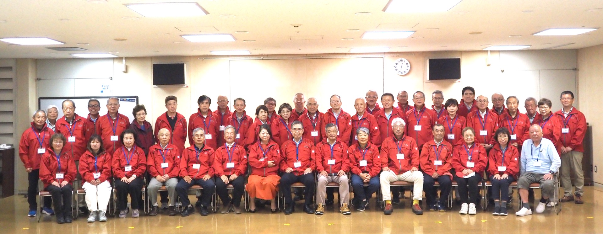 赤いジャケットの男女が会議室で集合写真、全員が名札付きで整列している。